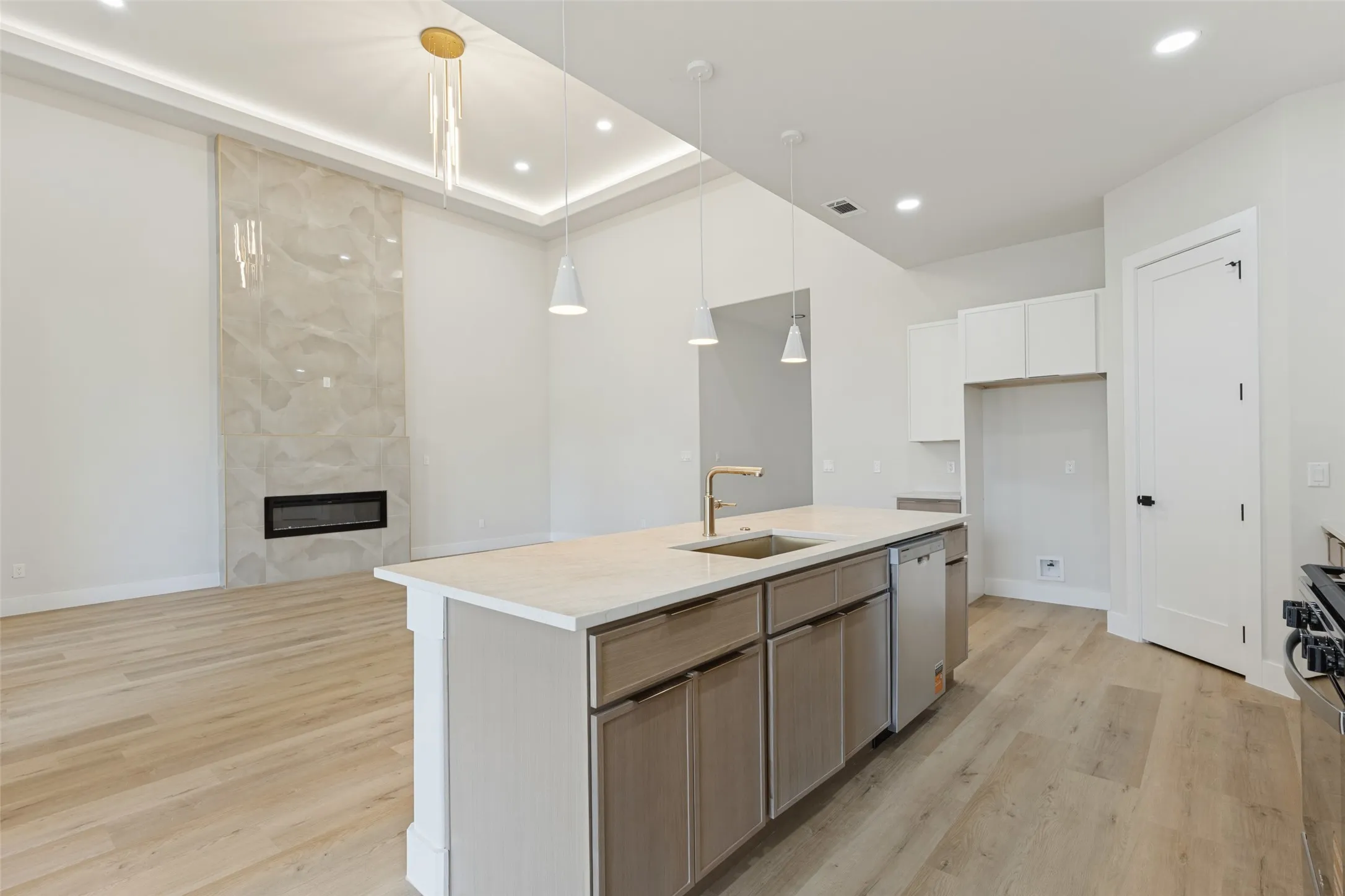 Spacious open-concept layout featuring wood-finish flooring, a contemporary kitchen island with a built-in sink, and a modern fireplace with a full-height stone-finish surround
