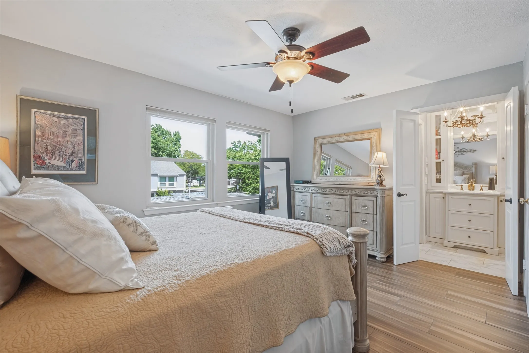 Wood-finish flooring, ceiling fan with integrated lighting, multiple windows, and an en-suite vanity area with a built-in cabinet and chandelier