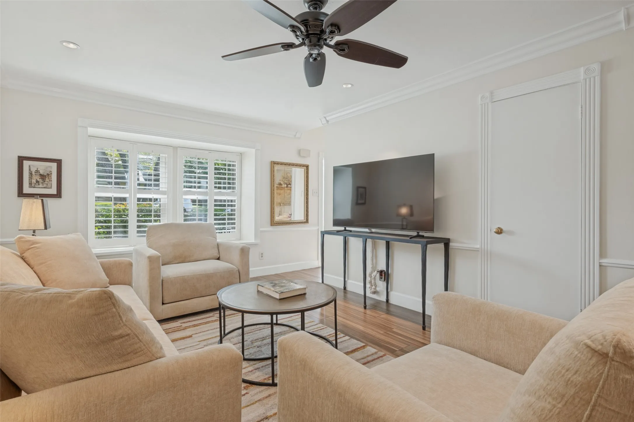 Crown molding, recessed lighting, ceiling fan, window with white plantation shutters, and wood-finish flooring