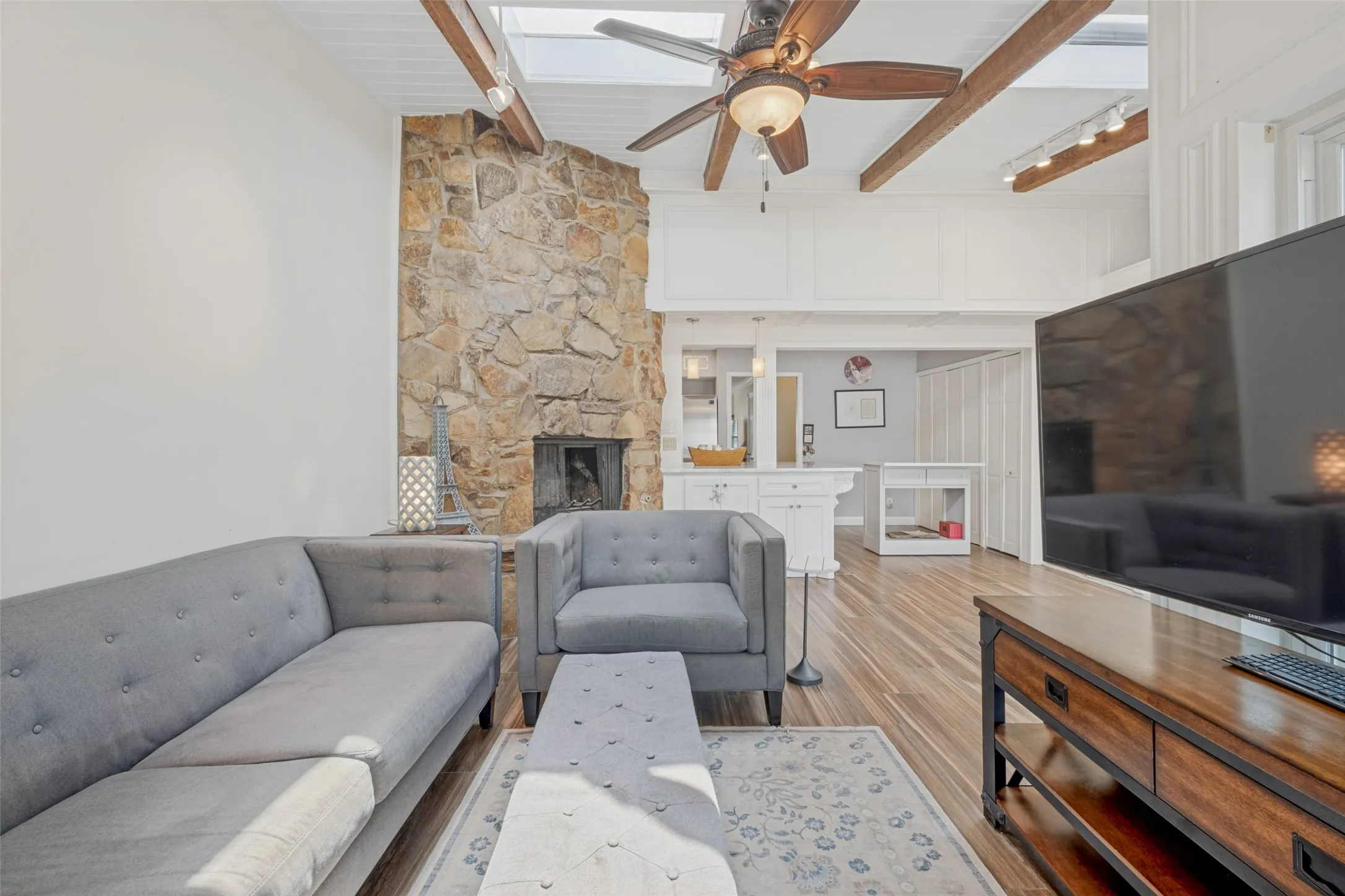 Vaulted ceiling with exposed wood beams, skylight, wood-finish flooring, stone fireplace, and ceiling fan