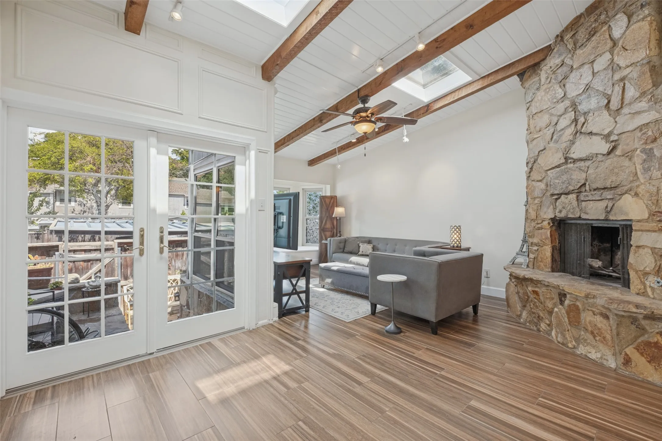 Vaulted ceiling with exposed wood beams, skylights, wood-finish flooring, stone fireplace, and French patio doors
