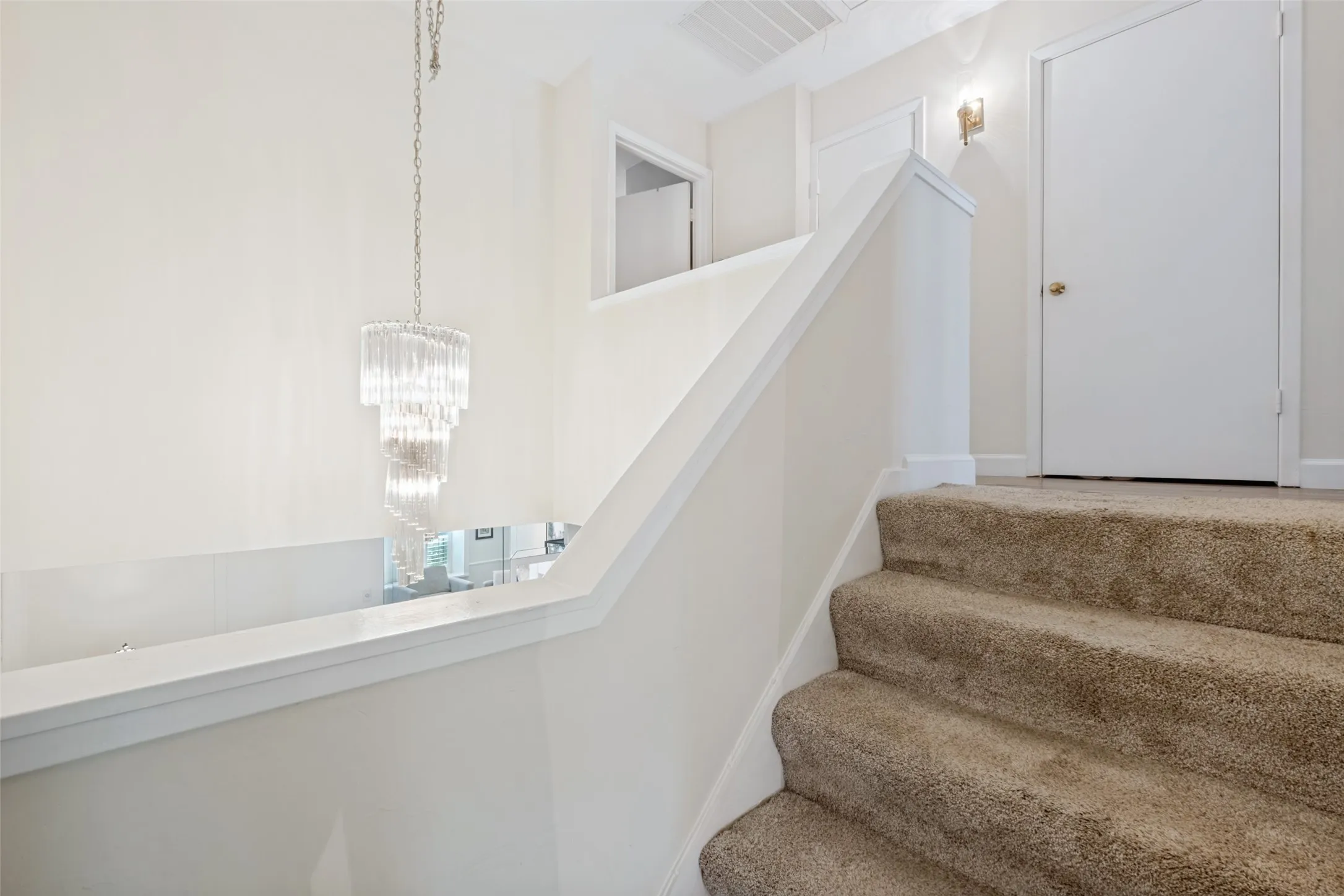 Carpeted staircase with white risers and handrail, featuring a wall-mounted sconce and a tiered crystal chandelier