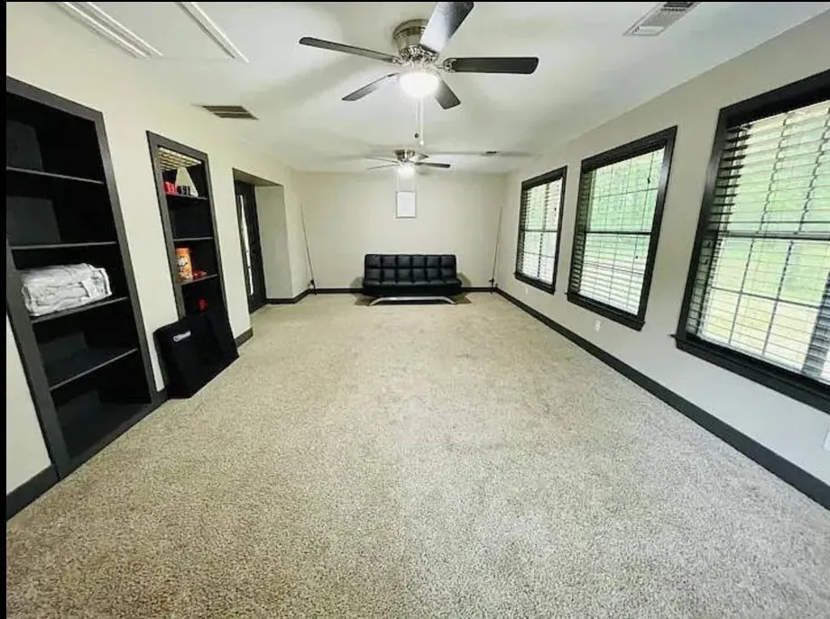 Spacious room featuring light-toned carpeting, multiple windows with blinds, built-in shelving, and ceiling fans