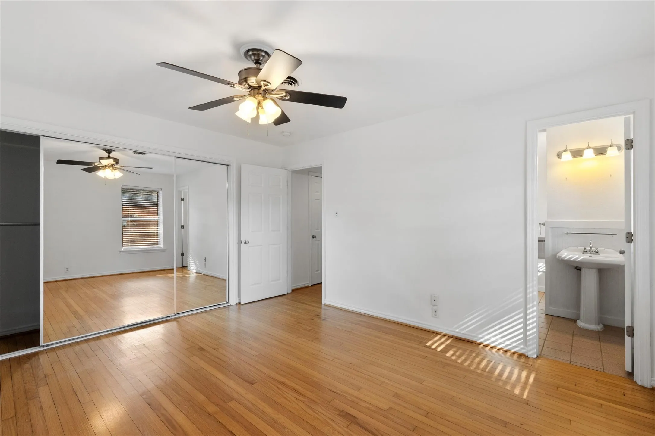 Wood-finish flooring, mirrored closet doors, a ceiling fan with integrated lighting, a pedestal sink, and wall-mounted vanity lighting