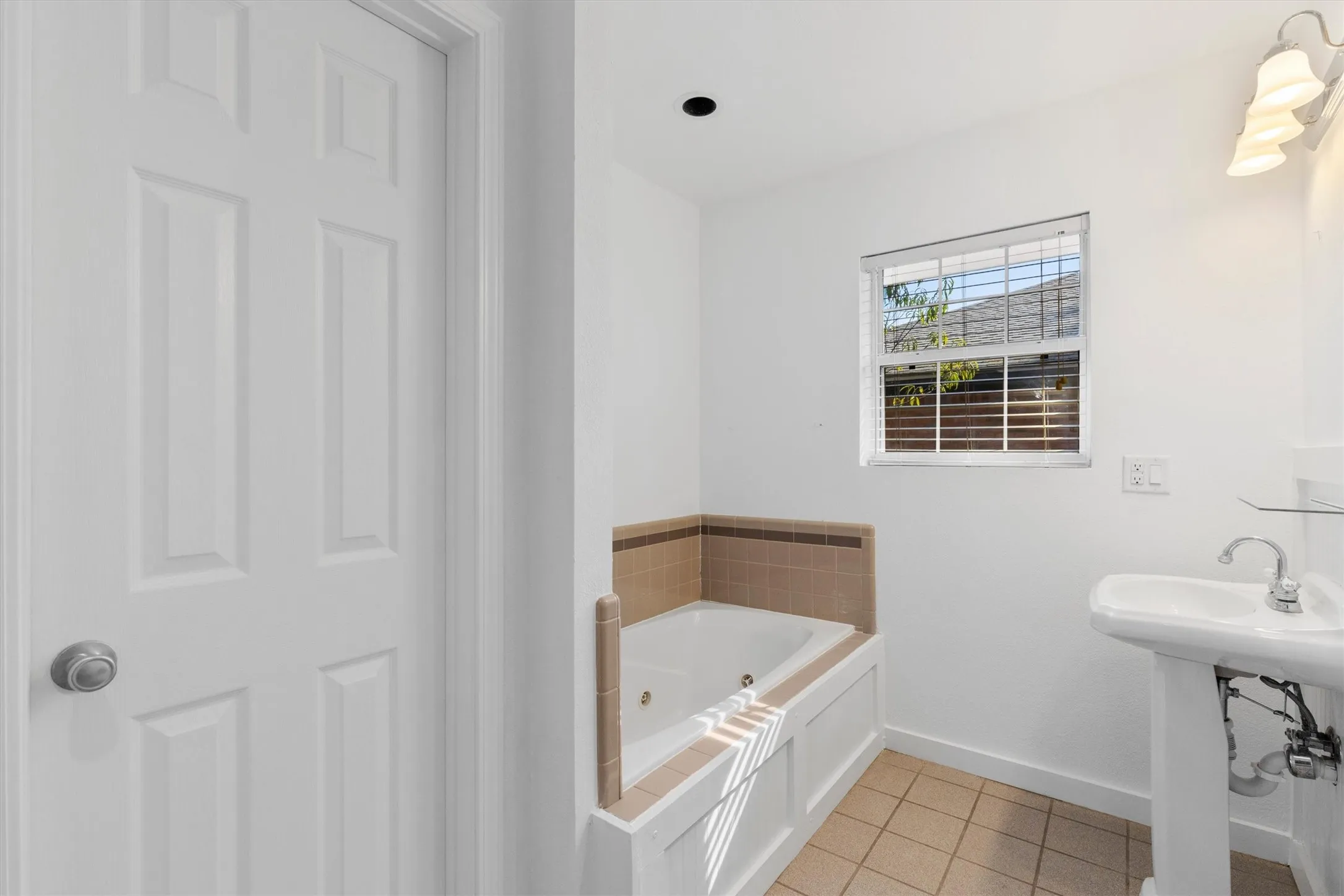 White six-panel door, built-in jetted tub with tiled surround, single window with blinds, pedestal sink with chrome faucet, and tiled flooring