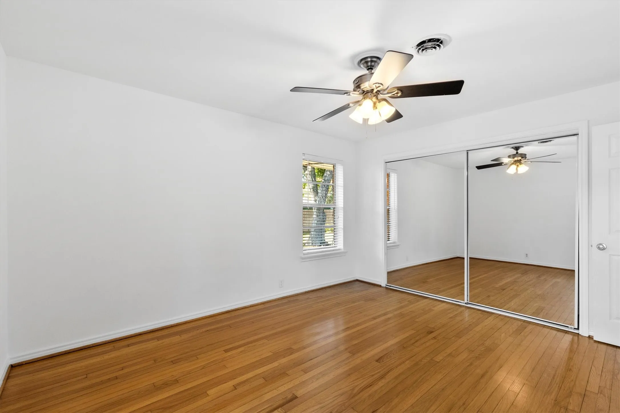 Hardwood flooring, mirrored closet doors, ceiling fan with integrated lighting, and a single window with horizontal blinds