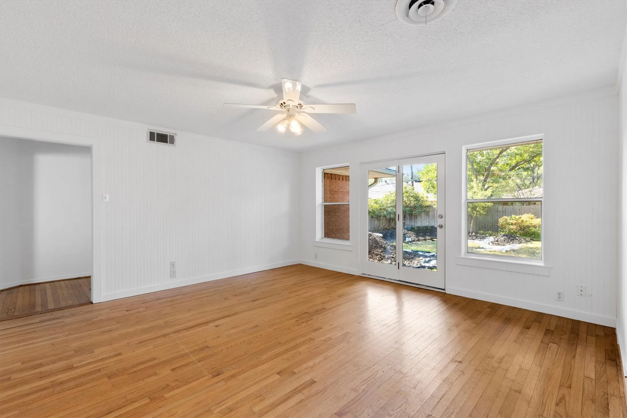 Hardwood flooring, white vertical plank wall panels, ceiling fan, double pane glass doors, and multiple windows