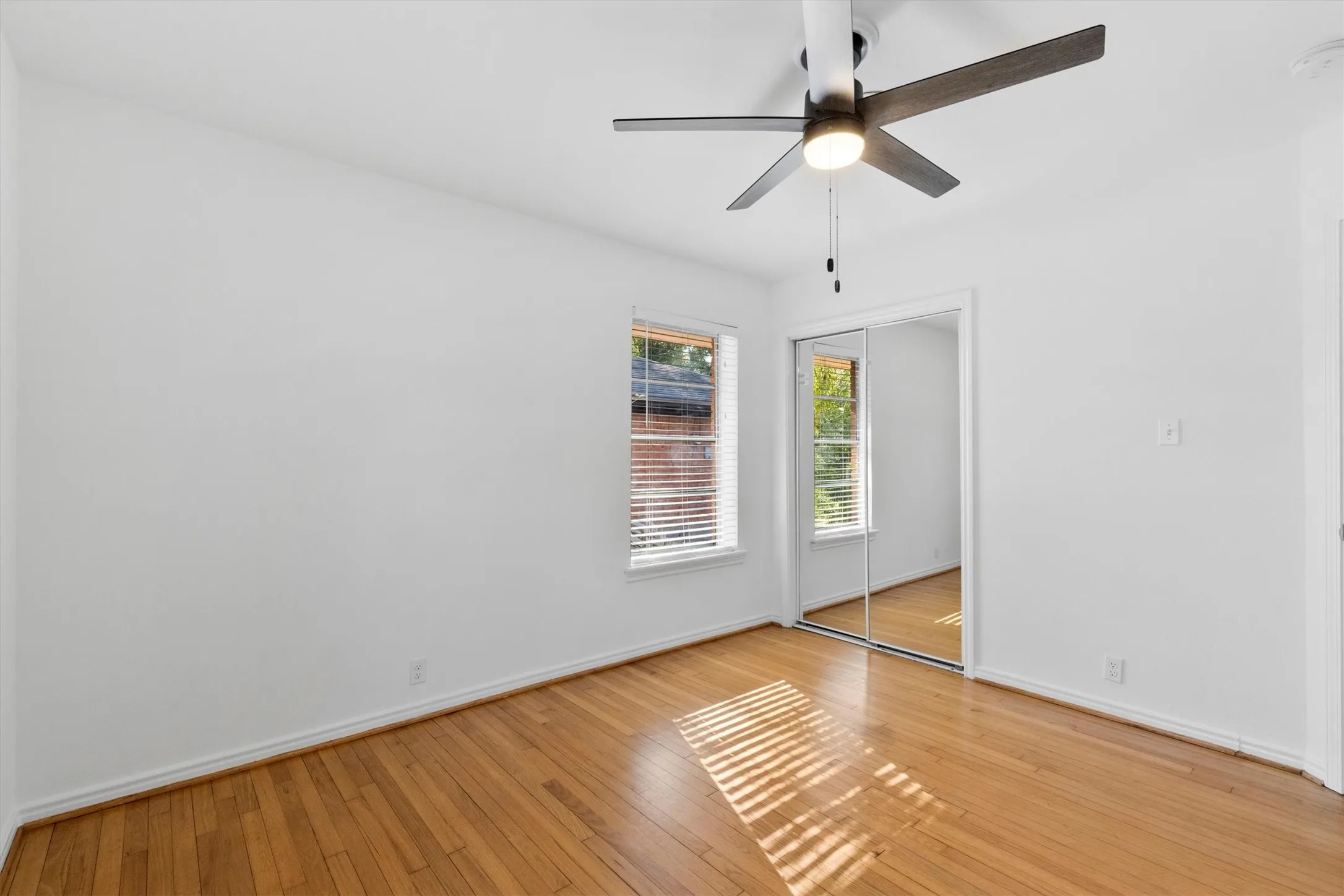 Hardwood flooring, white walls, window with blinds, mirrored sliding closet doors, and a ceiling fan with integrated lighting