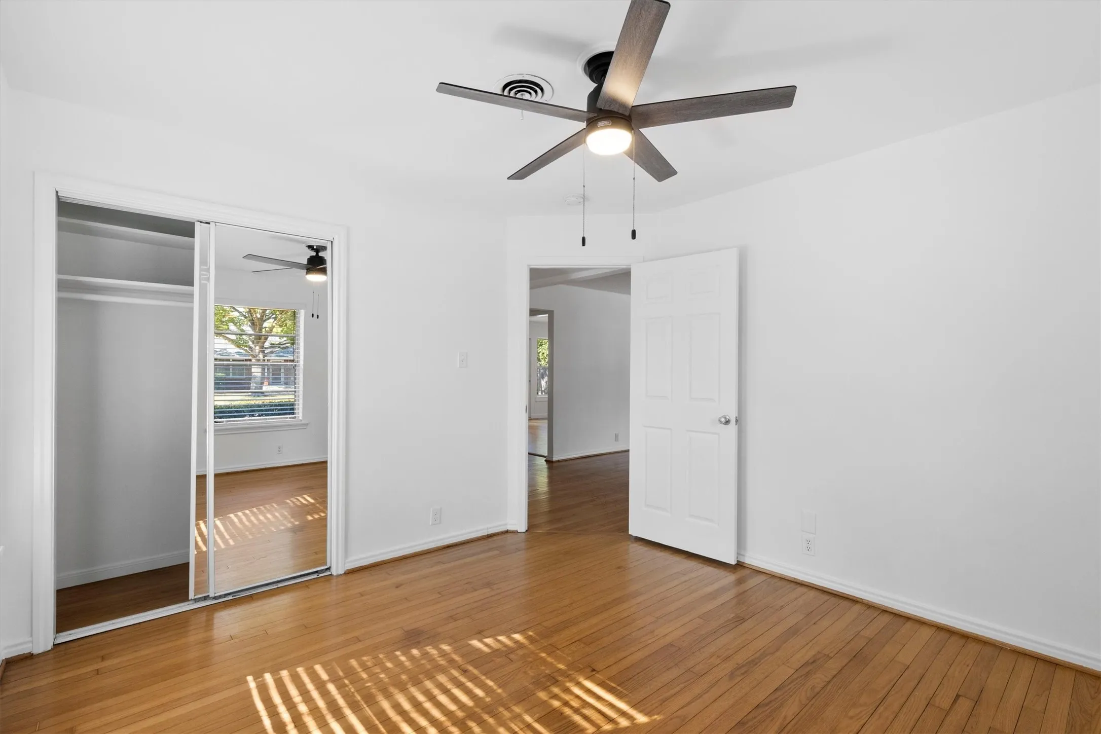 Hardwood flooring with a mirrored sliding closet door, white walls, and an overhead ceiling fan with lighting