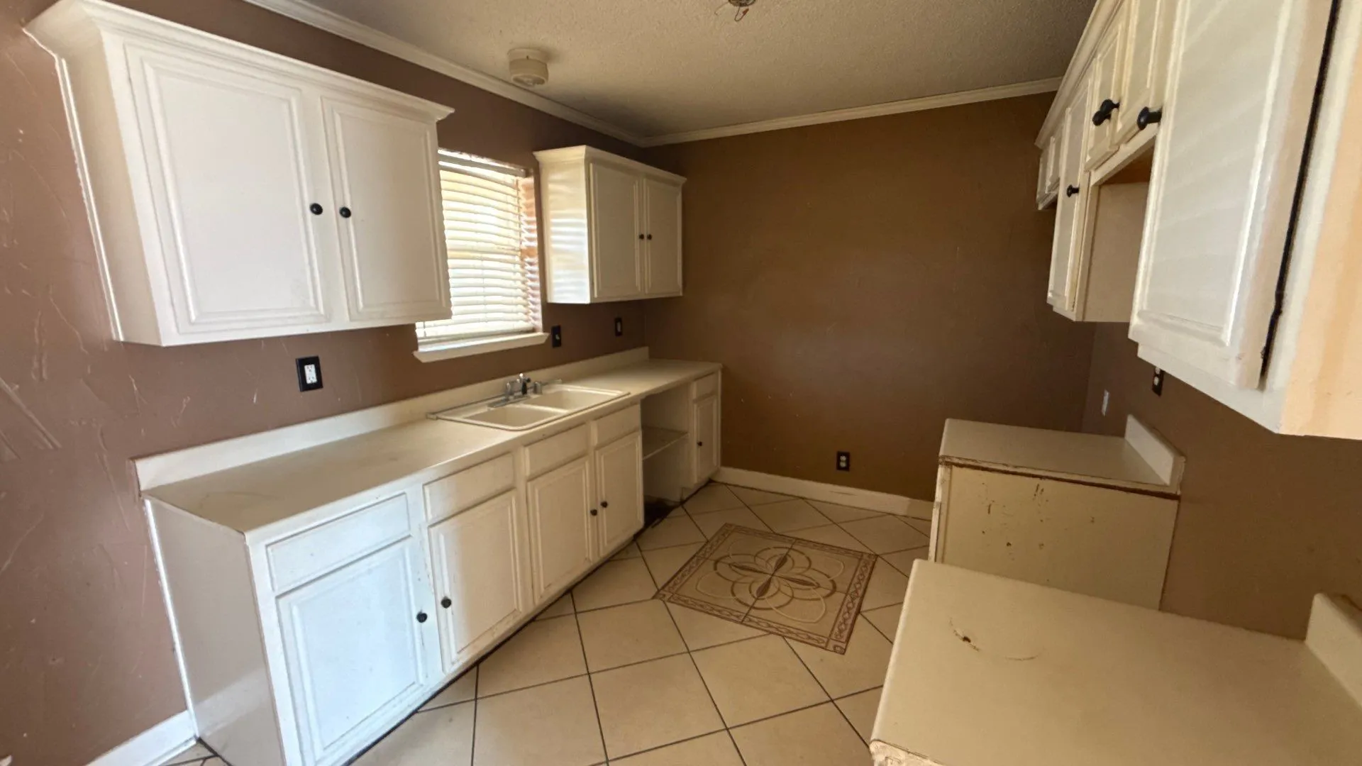 White shaker-style cabinetry with dark hardware, dual basin stainless steel sink, light-toned countertops, and tiled flooring with decorative inlay