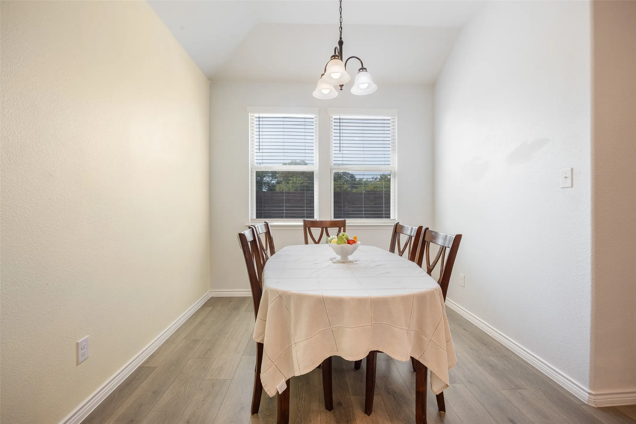 Vaulted ceiling with an attached chandelier, two windows with blinds, and wood-finish flooring