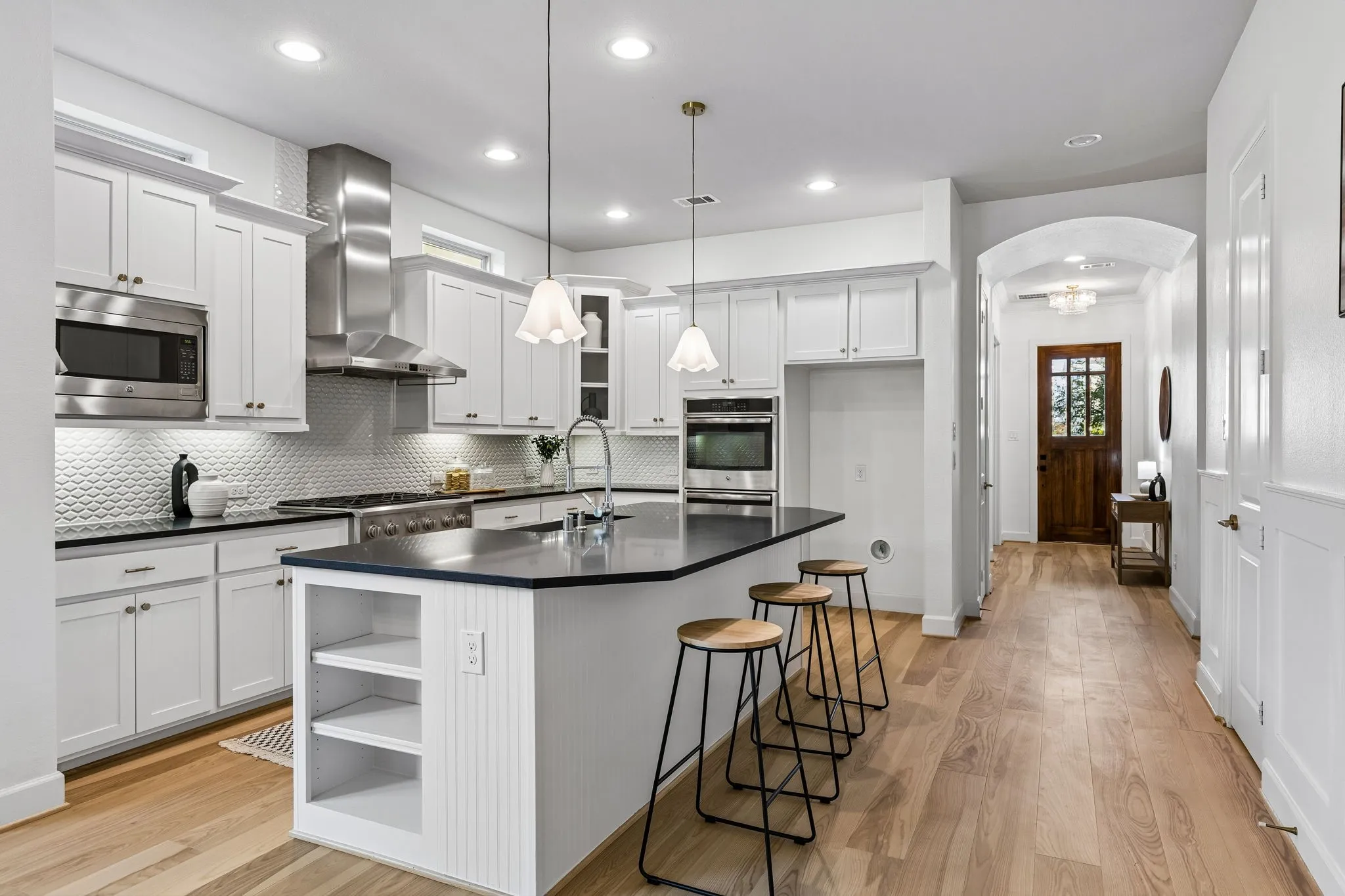Kitchen featuring white shaker-style cabinetry, stainless steel appliances, dark countertops, a large island with shelving, and wood-finish flooring