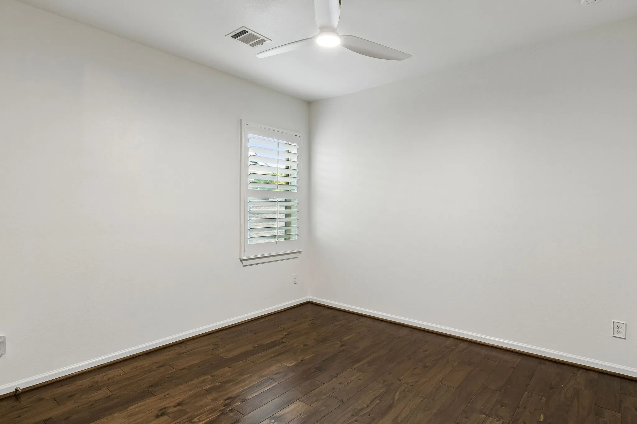 Wood-finish flooring, white walls, window with plantation shutters, and a ceiling fan with integrated lighting