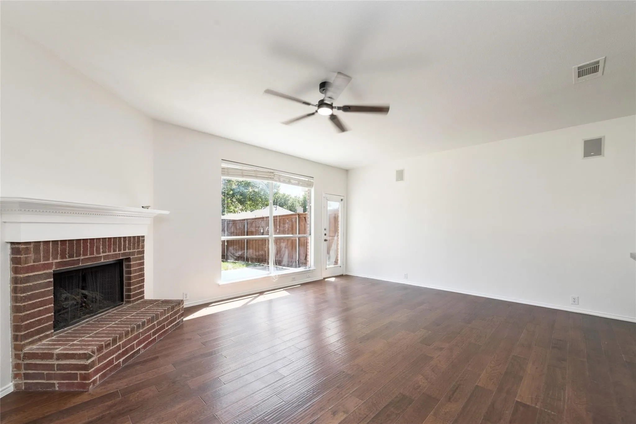 Vaulted ceiling with a ceiling fan, brick fireplace with mantel, wood-finish flooring, and a large window with a backyard view