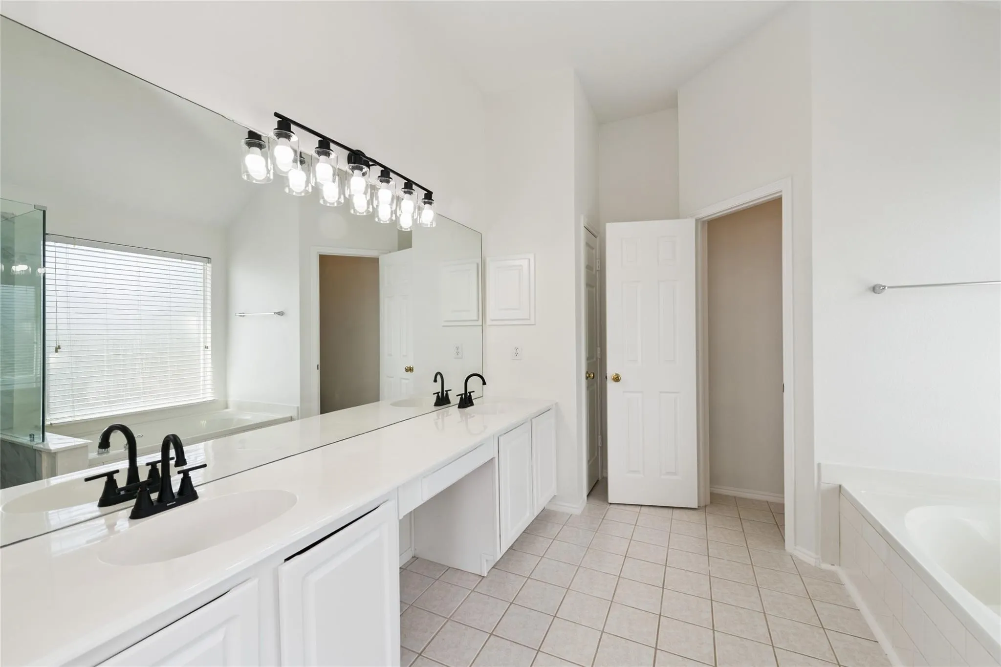Double vanity with undermount sinks and matte black faucets, extensive counter space, full-width wall mirror, and a built-in tub with a window
