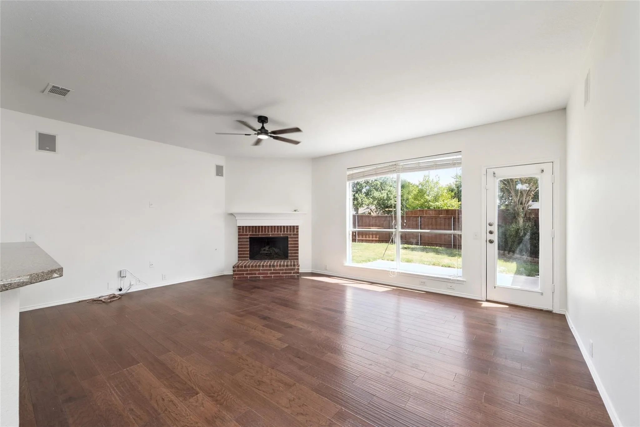 Spacious room featuring wood-finish flooring, a brick fireplace with mantel, a ceiling fan, and backyard access via a glass-paneled door