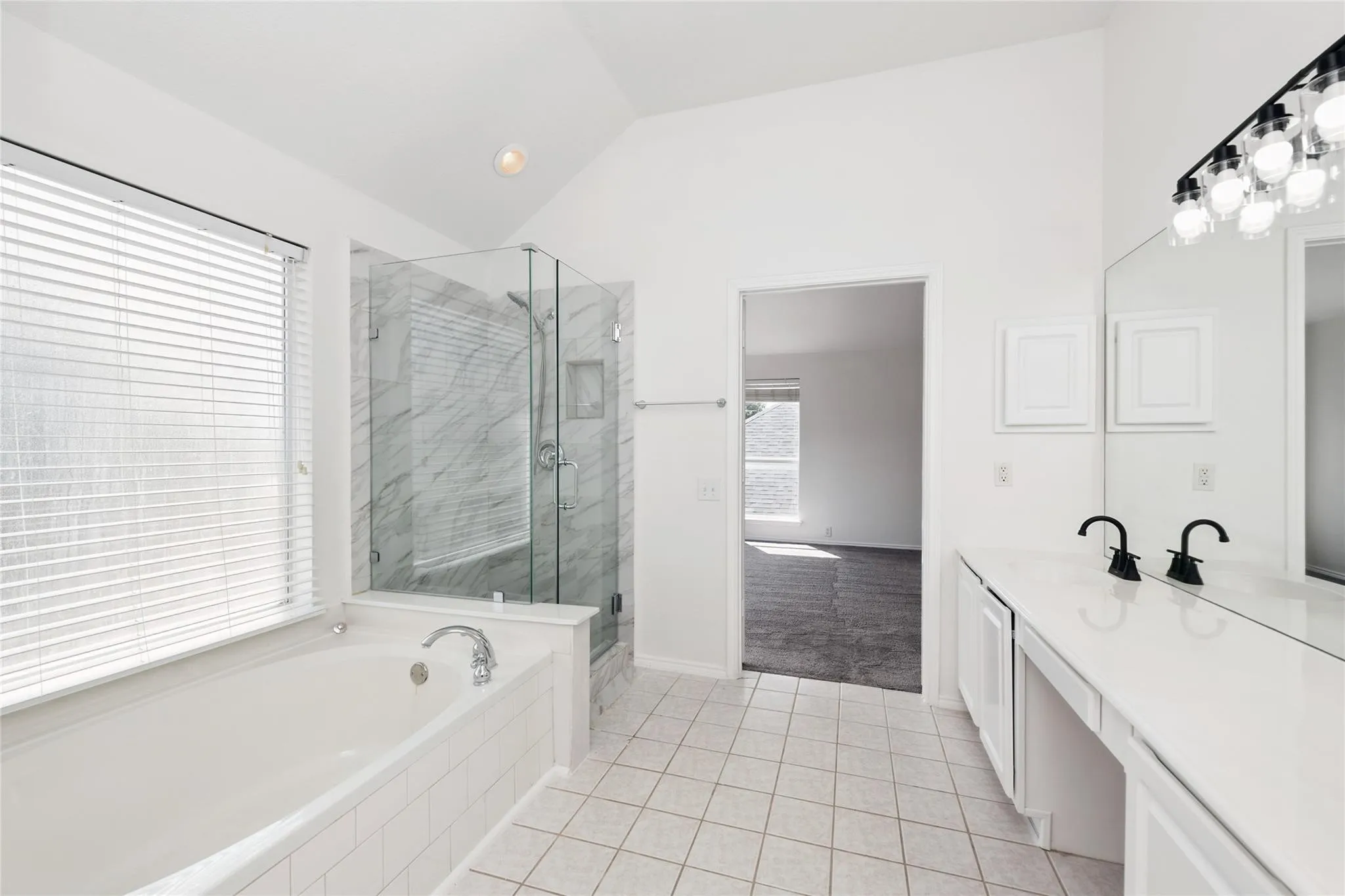 Soaking tub, glass-enclosed shower with tiled walls, dual vanity with black fixtures, vaulted ceiling, and tiled flooring