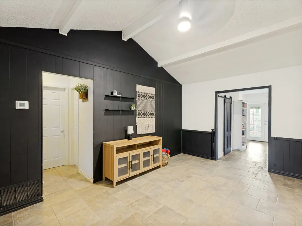 Vaulted ceilings with exposed beams, plank accent wall, neutral-toned tile flooring, and a sliding barn door