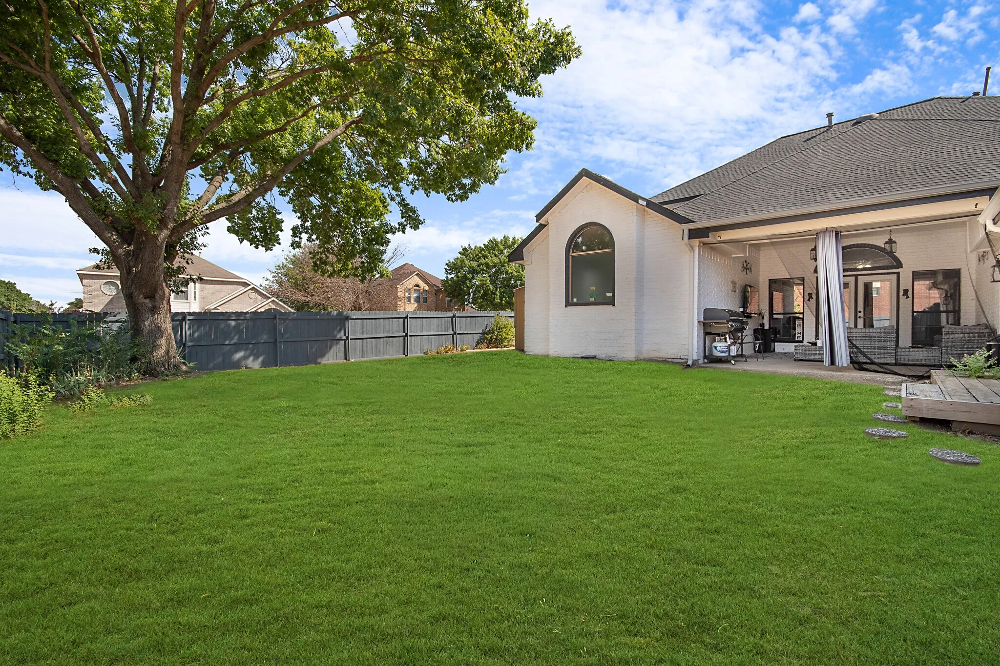 Deep fenced back yard under a mature shade tree, with the covered patio running along the back of the house.