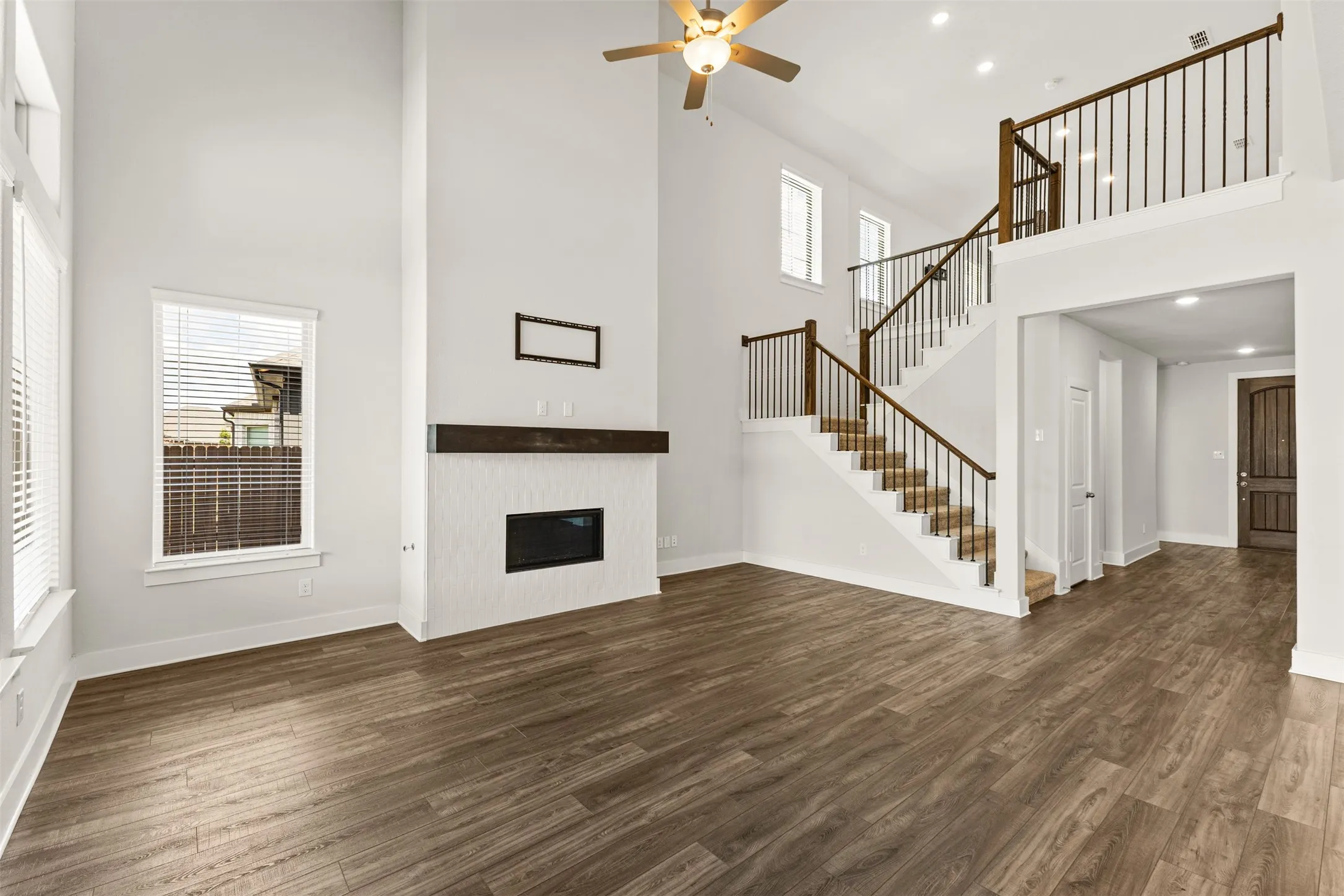 Soaring ceilings with recessed lighting, wood-finish flooring, a built-in electric fireplace with a dark wood mantel, and a multi-level staircase with dark wood handrails and wrought iron balusters