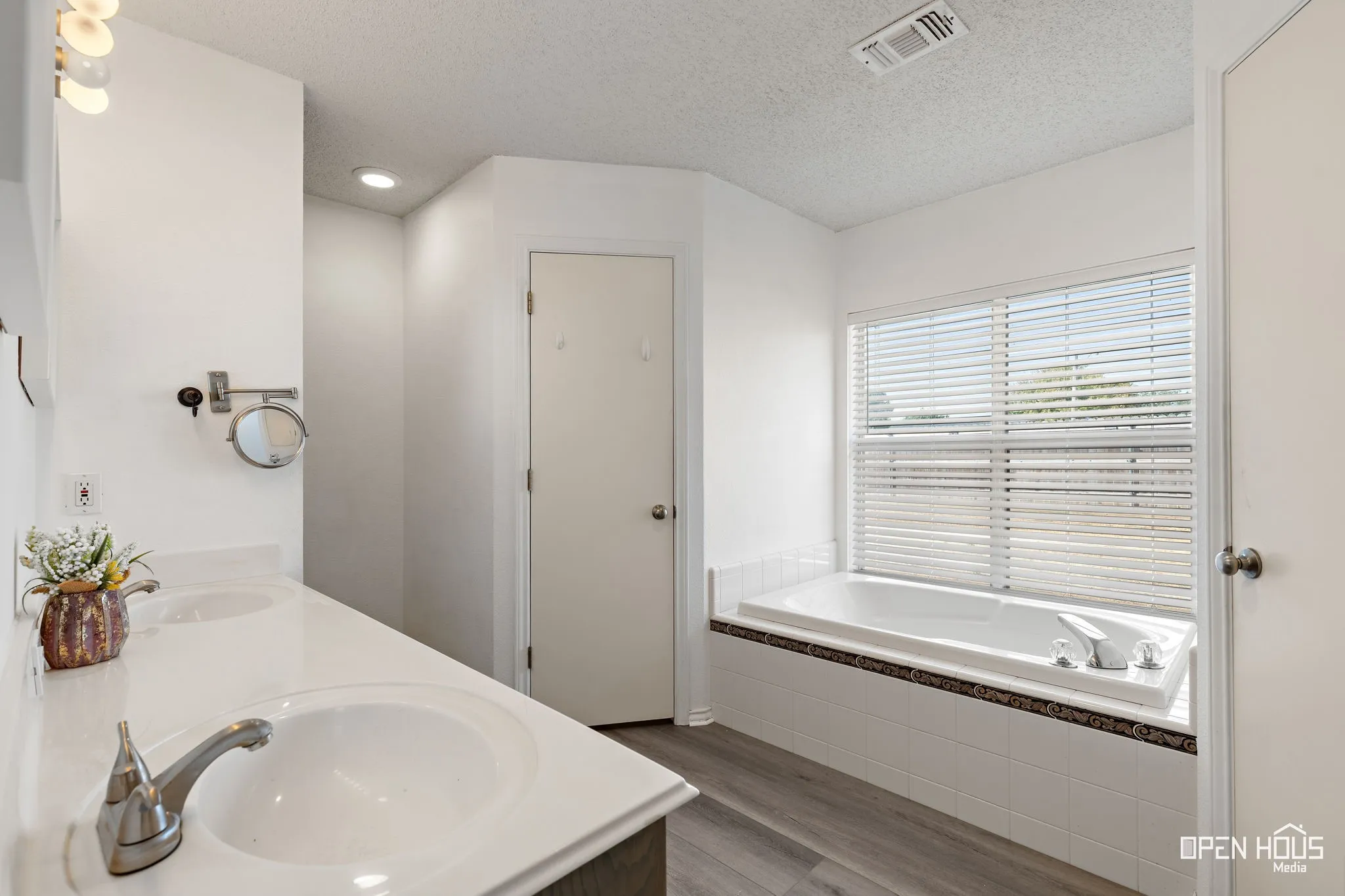 Dual vanity with integrated sinks, brushed nickel fixtures, a built-in soaking tub, a window with blinds, and wood-finish flooring