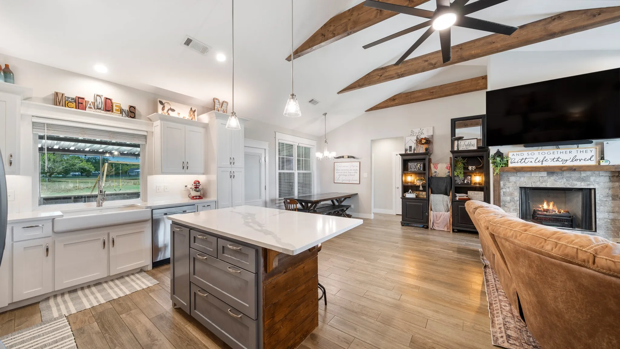 Vaulted ceiling with exposed wood beams, wood-finish flooring, central kitchen island with a light-toned countertop, white cabinetry, and a stone-clad fireplace