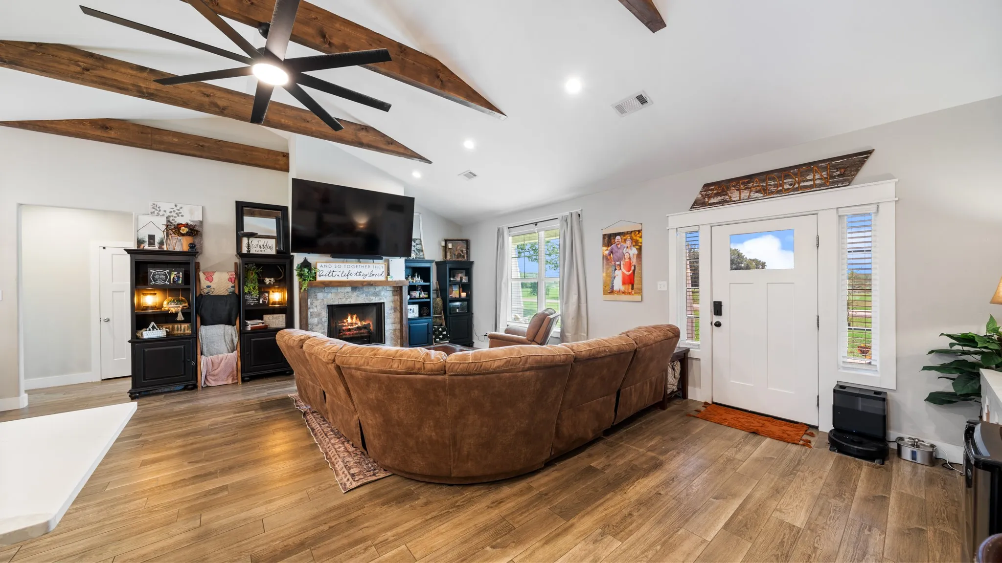 Vaulted ceiling with wood beams, wood-finish flooring, stone-faced fireplace with mantle, recessed lighting, and a front entry door with transom and sidelights
