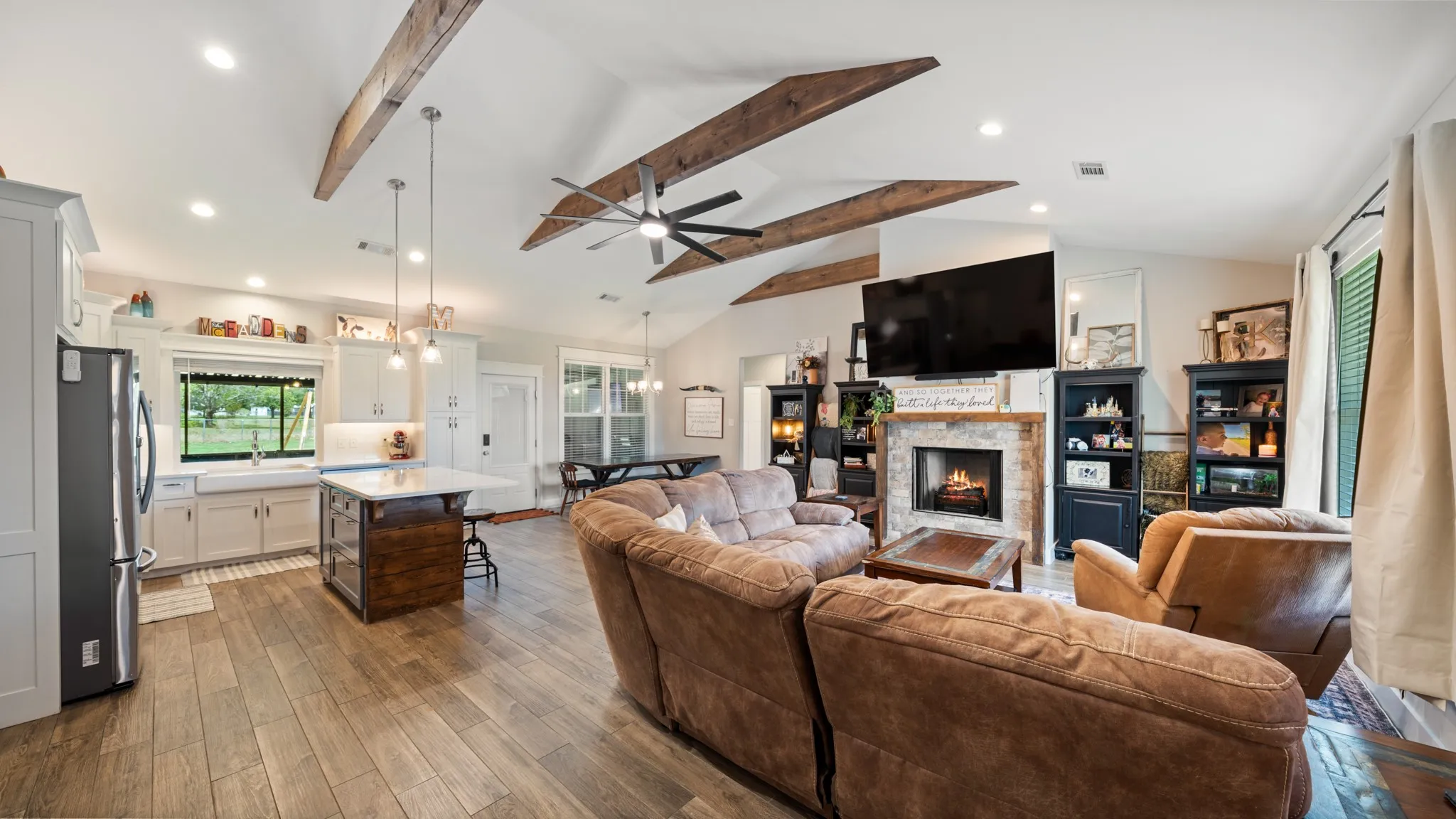 Vaulted ceiling with exposed wood beams, stone fireplace, wood-finish flooring, white cabinetry, and a kitchen island with a light-toned countertop