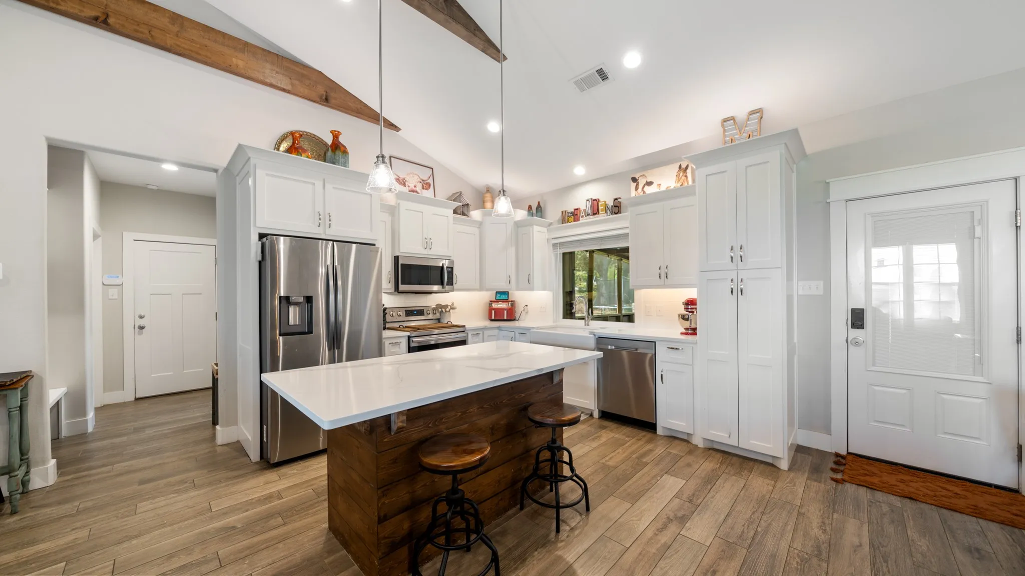Vaulted ceiling with exposed wood beams, bright white cabinetry, stainless steel appliances, wood-finish flooring, and a central island with a solid surface countertop