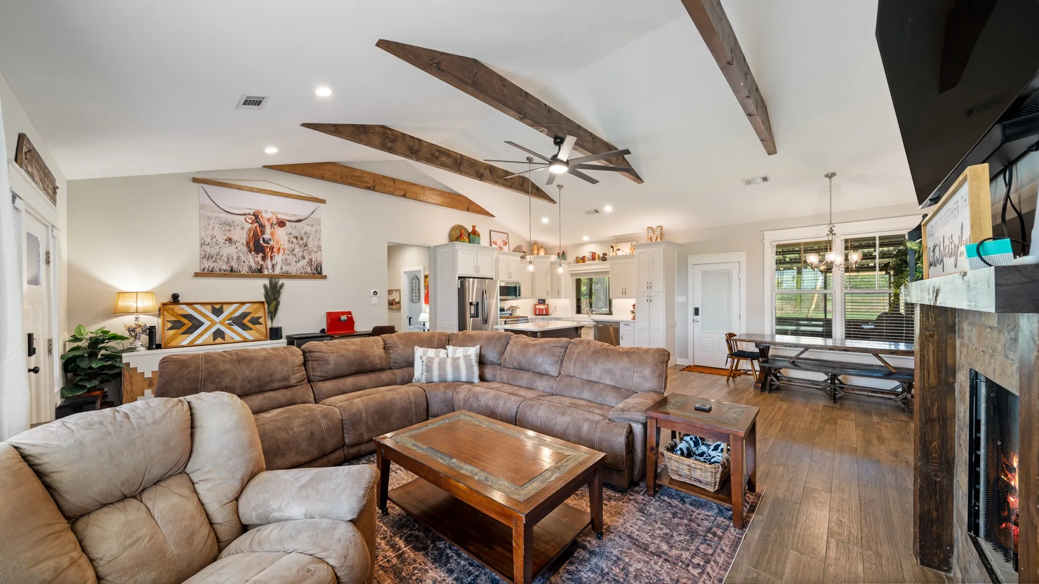 Vaulted ceiling with exposed wood beams, wood-finish flooring, fireplace with stone surround, recessed lighting, and a ceiling fan