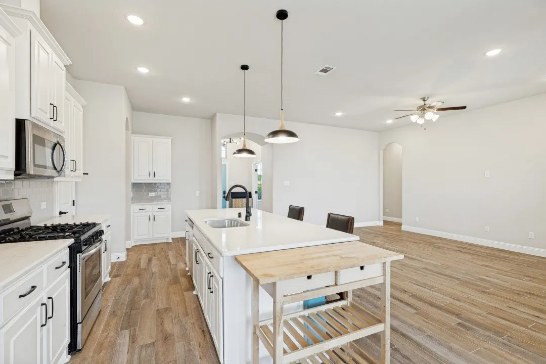 Kitchen featuring white cabinetry, stainless steel appliances, a center island with sink, pendant lighting, and wood-finish flooring also notice the wide-open living room. And recess lights