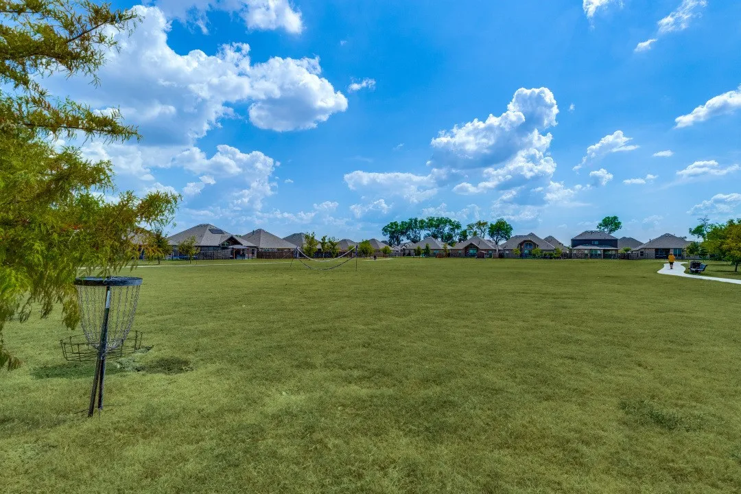 Expansive green space with disc golf basket, bordered by residences with varied rooflines and mature trees, featuring a paved pathway