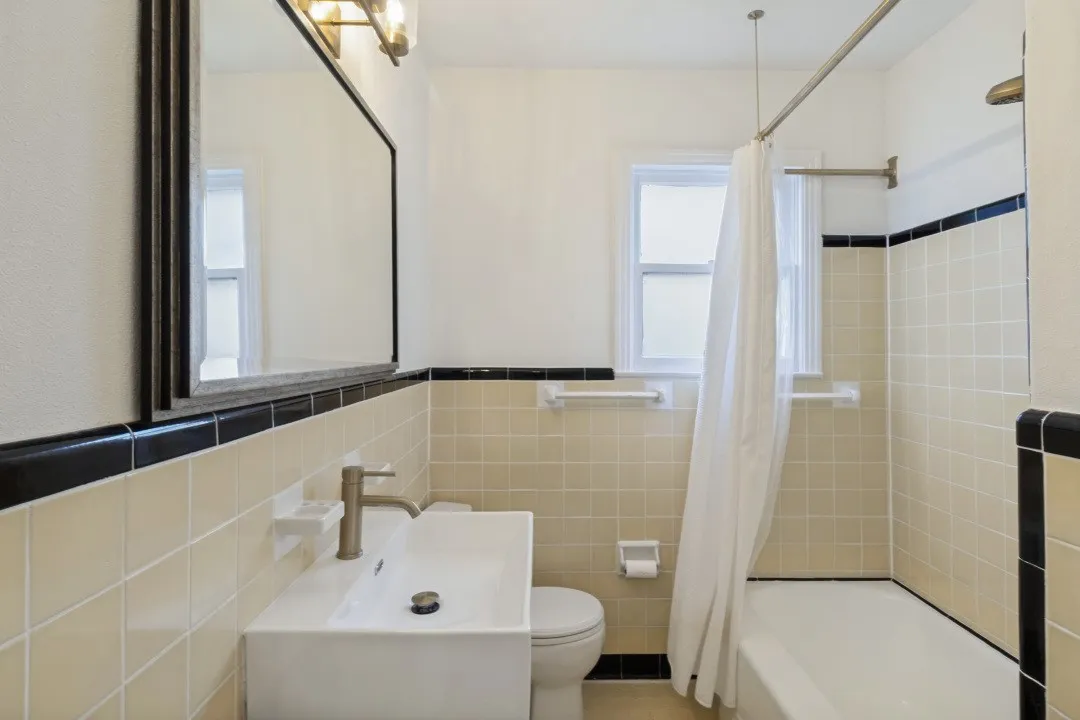Rectangular wall mirror, vanity with a white vessel sink, single-handle faucet, window with privacy glass, and tiled shower/tub combination