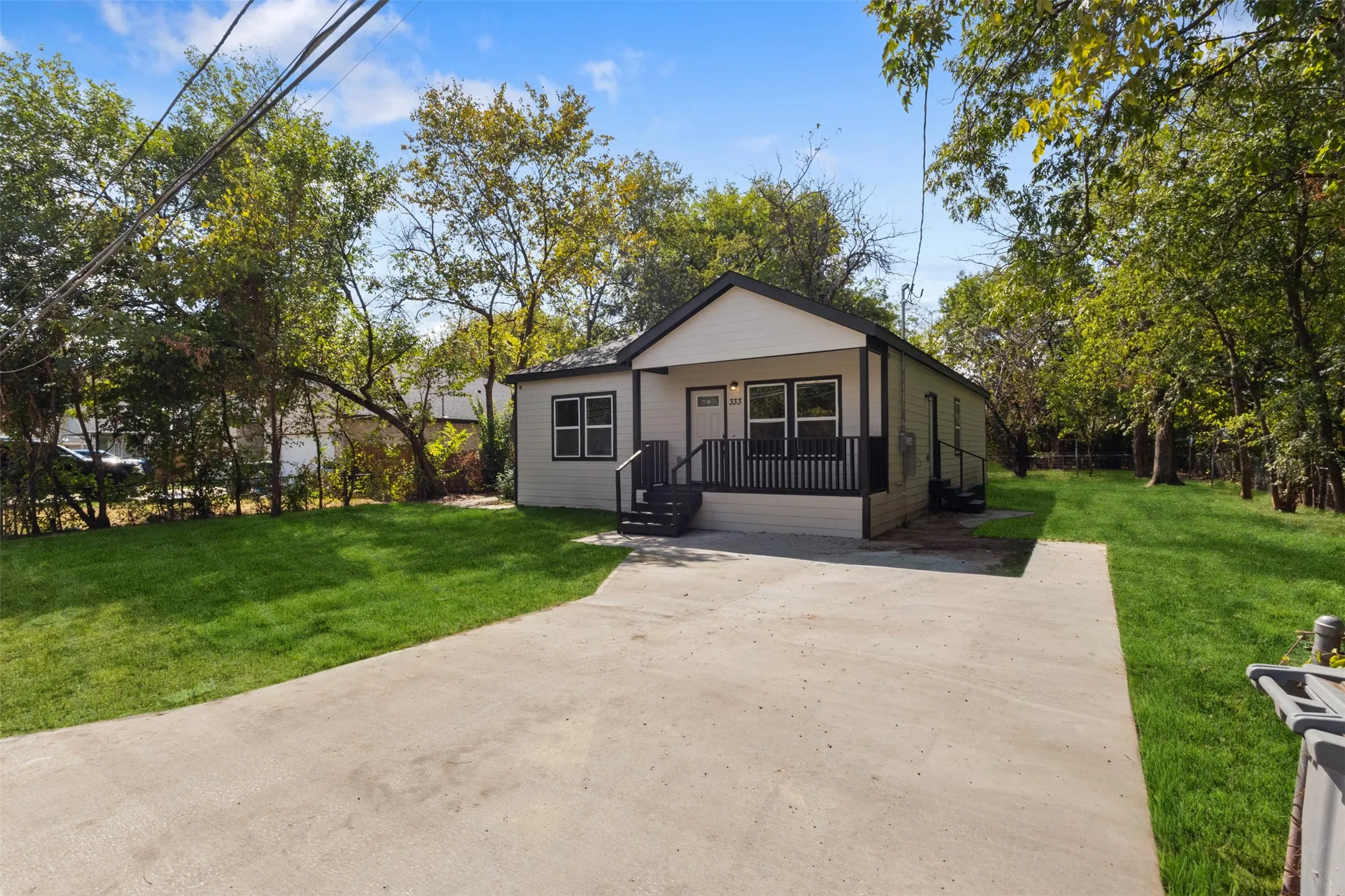 Contemporary exterior with light siding and dark trim, featuring a covered front porch with dark railing, a concrete driveway, and surrounding mature trees