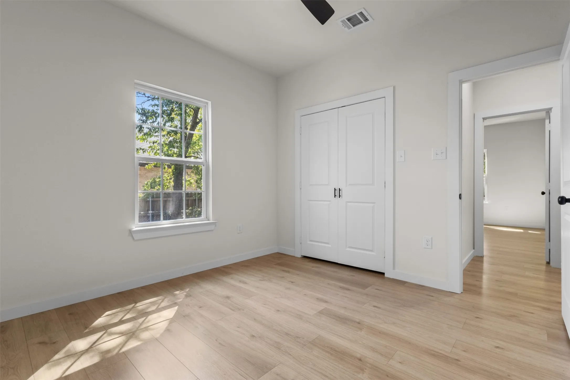 Wood-finish flooring, white walls, double-door closet, window with exterior foliage, and a ceiling fan