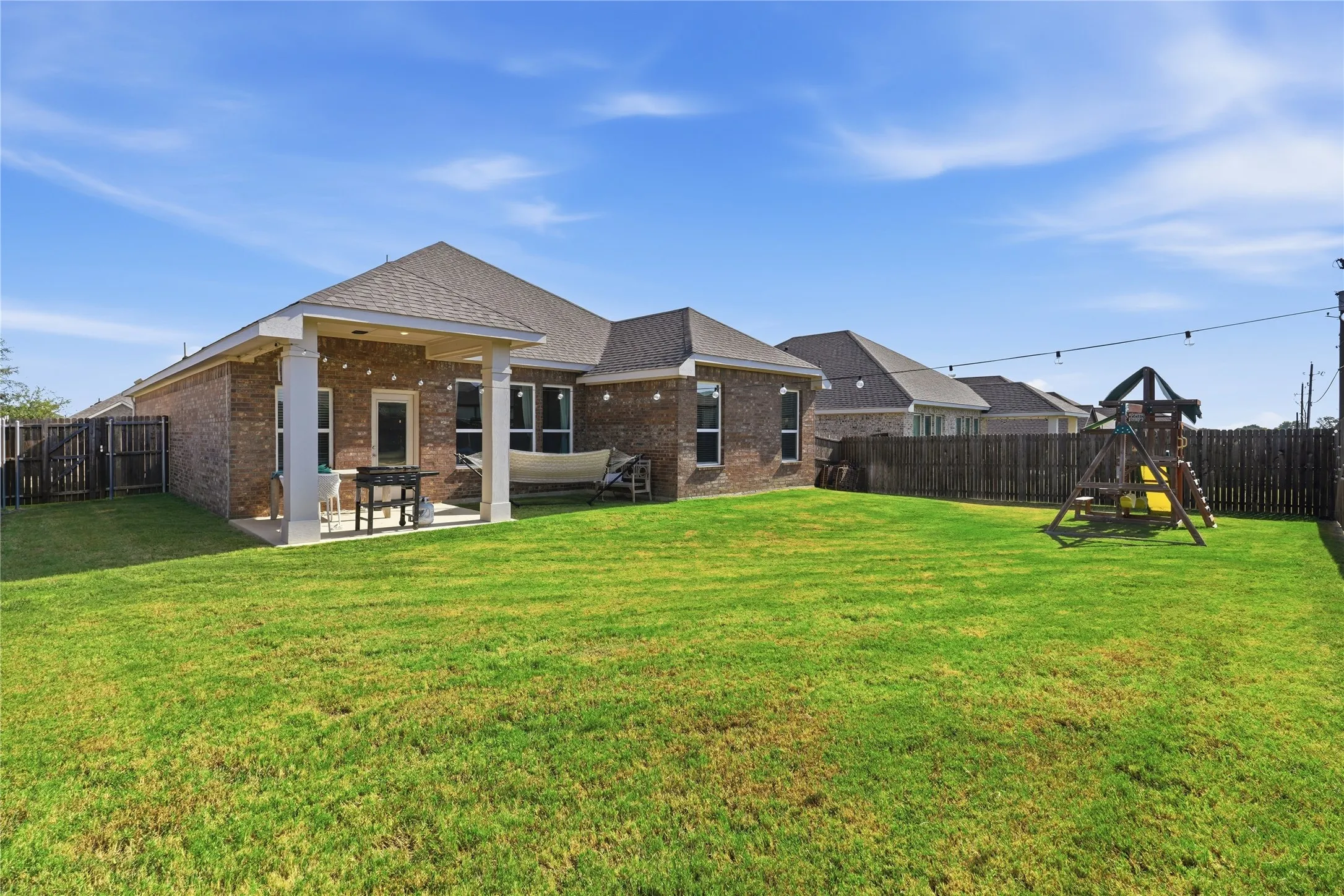 Expansive green lawn with a covered patio featuring square columns, a brick exterior, and a wooden privacy fence