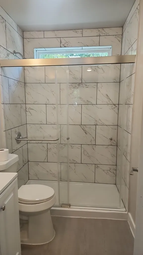 Tiled shower with a clear glass enclosure, overhead transom window, and a grab bar