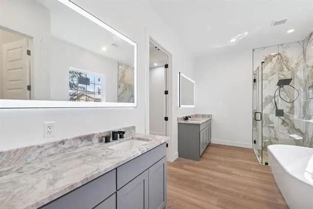 Dual vanities with stone countertops, wood-finish flooring, a freestanding bathtub, and a glass-enclosed shower