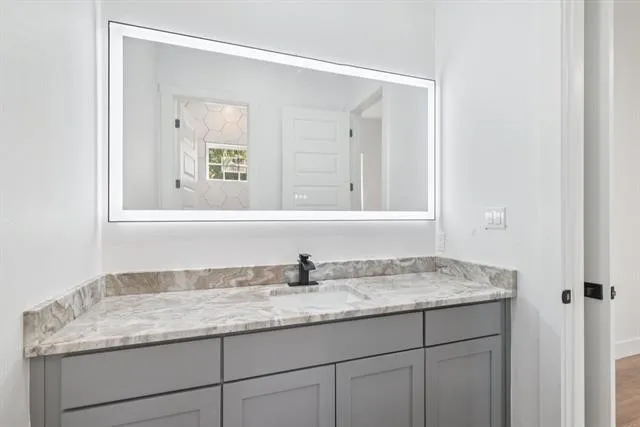 Illuminated rectangular vanity mirror over a gray cabinet with a built-in sink, stone-finish countertop, and matte black faucet