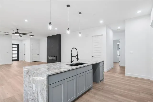 Central kitchen island with a stone countertop and integrated sink, gray cabinetry, recessed lighting, wood-finish flooring, and a modern fireplace