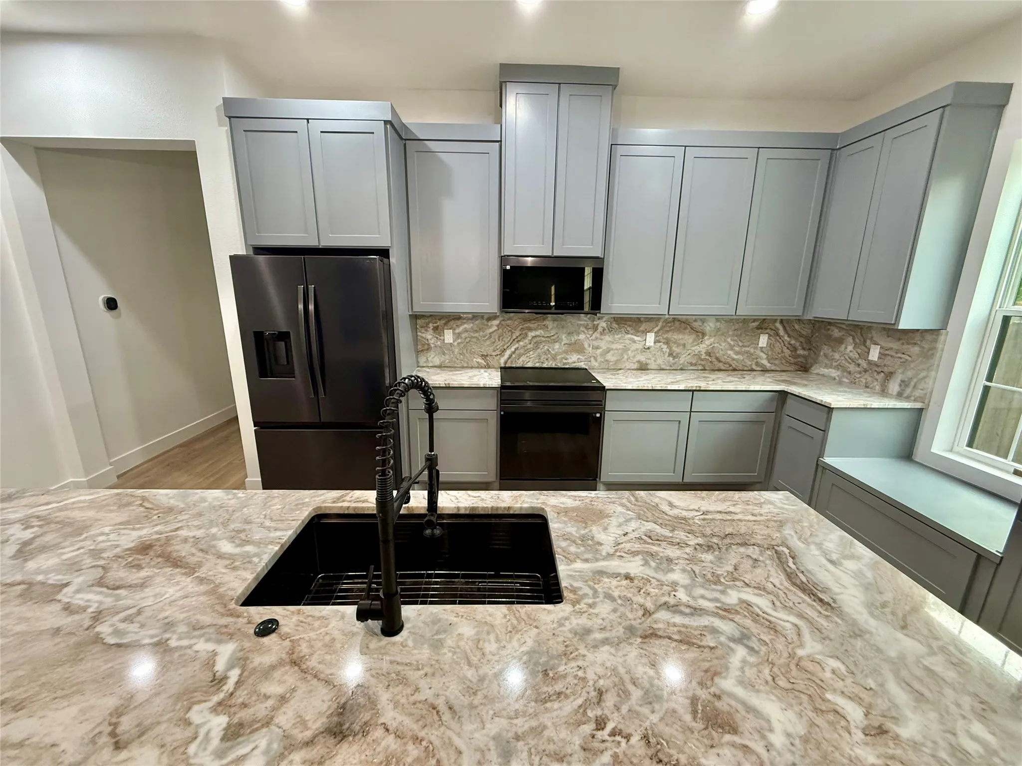 Kitchen featuring a large island with a black basin sink and pull-down faucet, light gray cabinetry, built-in stainless steel appliances, a window with a built-in bench, and patterned countertops with matching backsplash