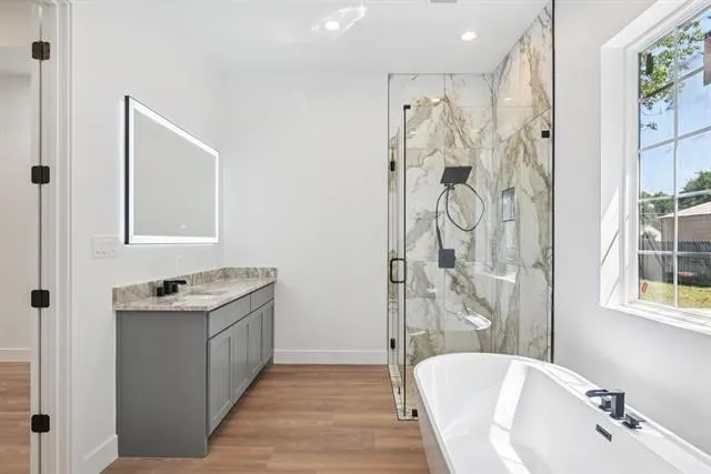 Freestanding soaking tub, walk-in shower with glass enclosure, single vanity with stone countertop, wood-finish flooring, and a large window