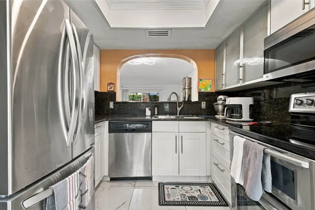 Stainless steel appliance suite, white shaker-style cabinetry, dark stone countertops, contemporary tile flooring, and an arched pass-through window