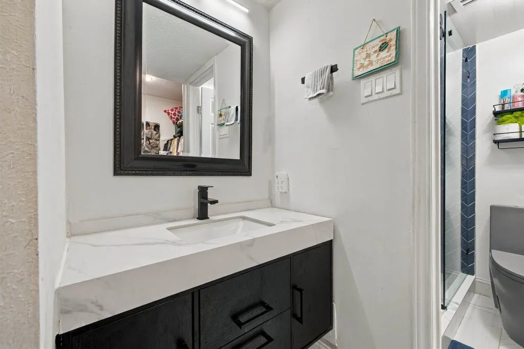 Vanity with integrated sink and matte black faucet, dark wood-finish cabinetry, framed mirror, and glass-enclosed shower with blue chevron tile accent