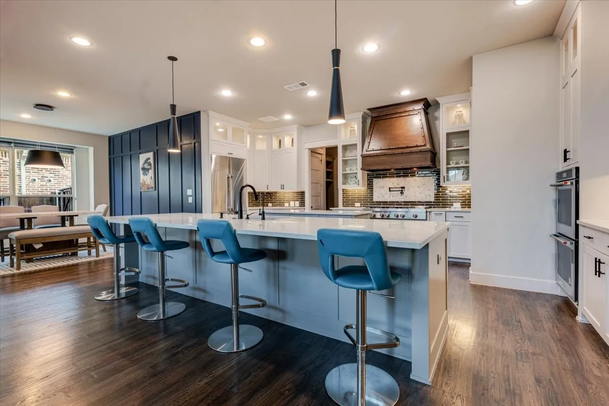 Kitchen featuring an expansive island with a light-toned countertop, wood-finish flooring, white cabinetry, a built-in double oven, and a decorative range hood