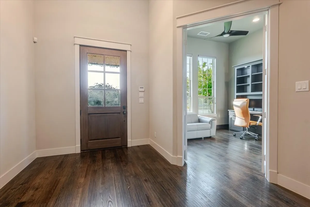 Wood-finish flooring throughout, solid wood entry door with glass panels, room with built-in shelving, windows providing natural light, and a ceiling fan