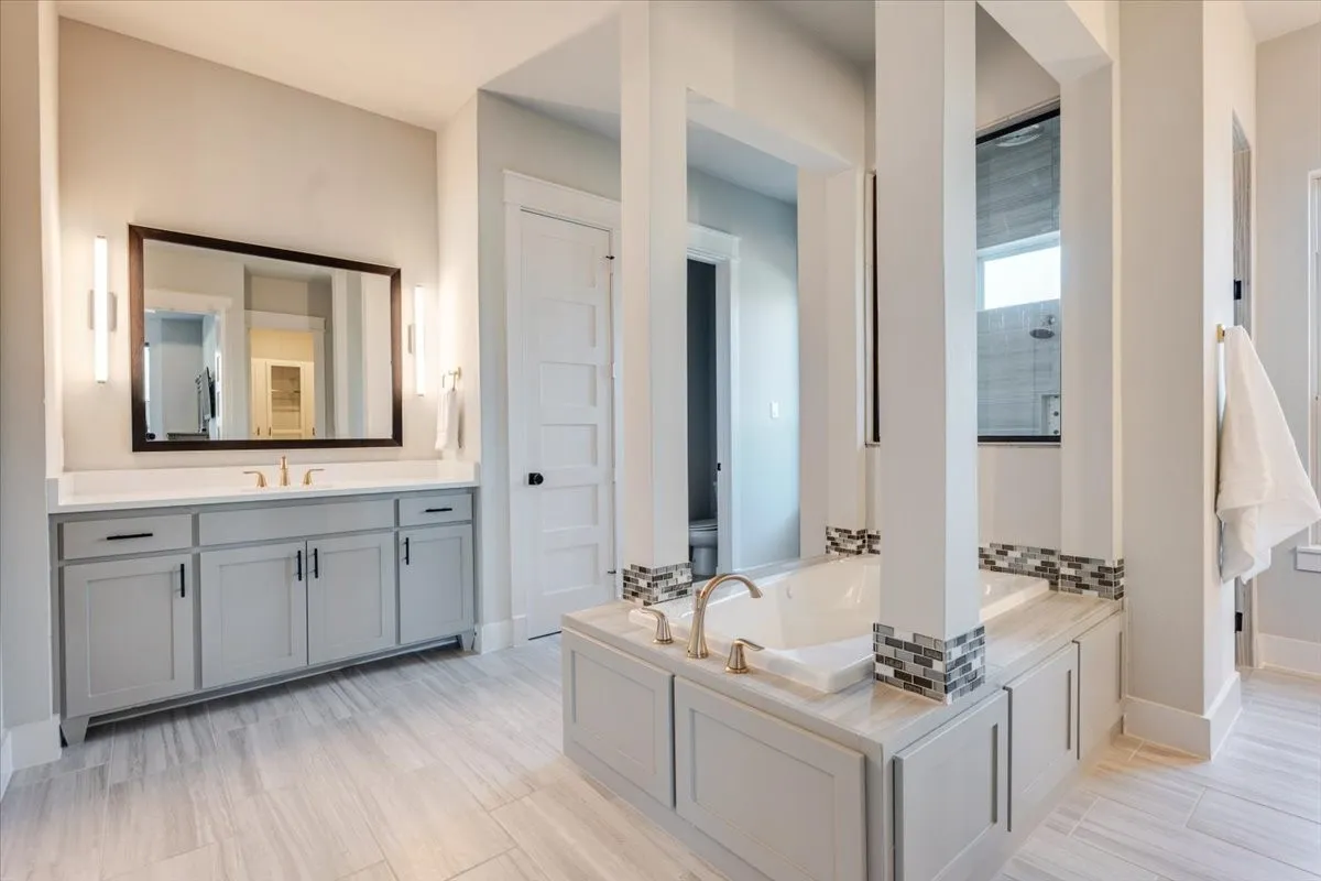 Wood-look tile flooring, built-in soaking tub with tile surround, dual vanity with shaker-style cabinetry, rectangular undermount sink, and wall-mounted sconce lighting