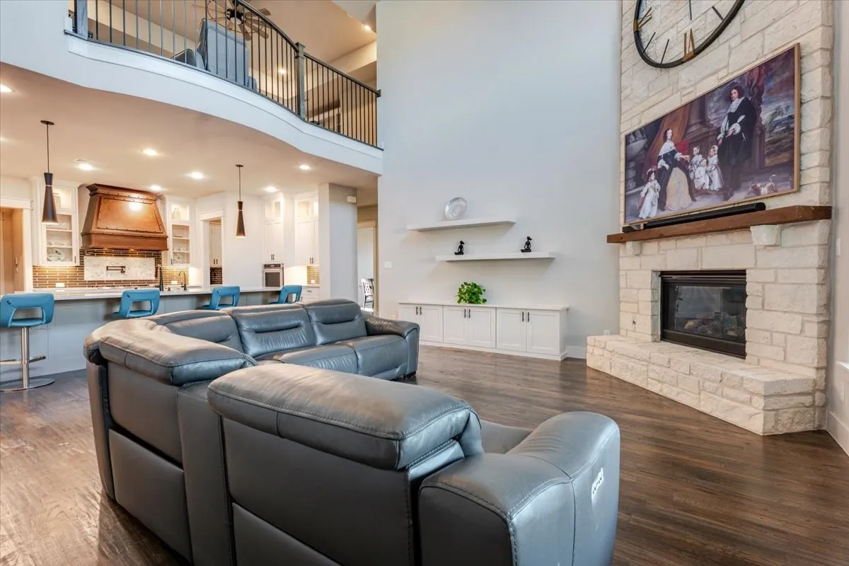 Two-story ceiling, stone-surround fireplace with wood mantel, wood-finish flooring, built-in cabinetry, and a second-story balcony with metal railing