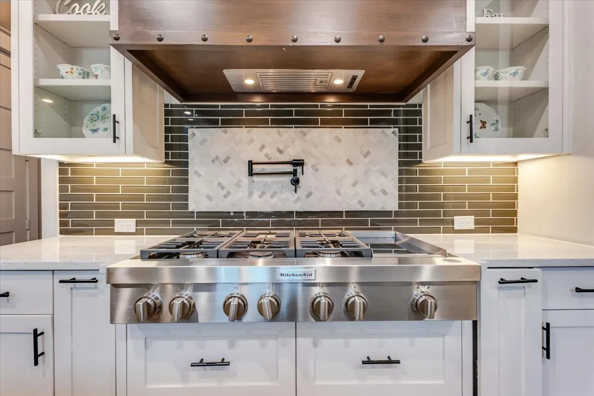 Stainless steel gas range with a wood-finish hood, solid surface countertops, glass-front upper cabinetry, and a subway tile backsplash