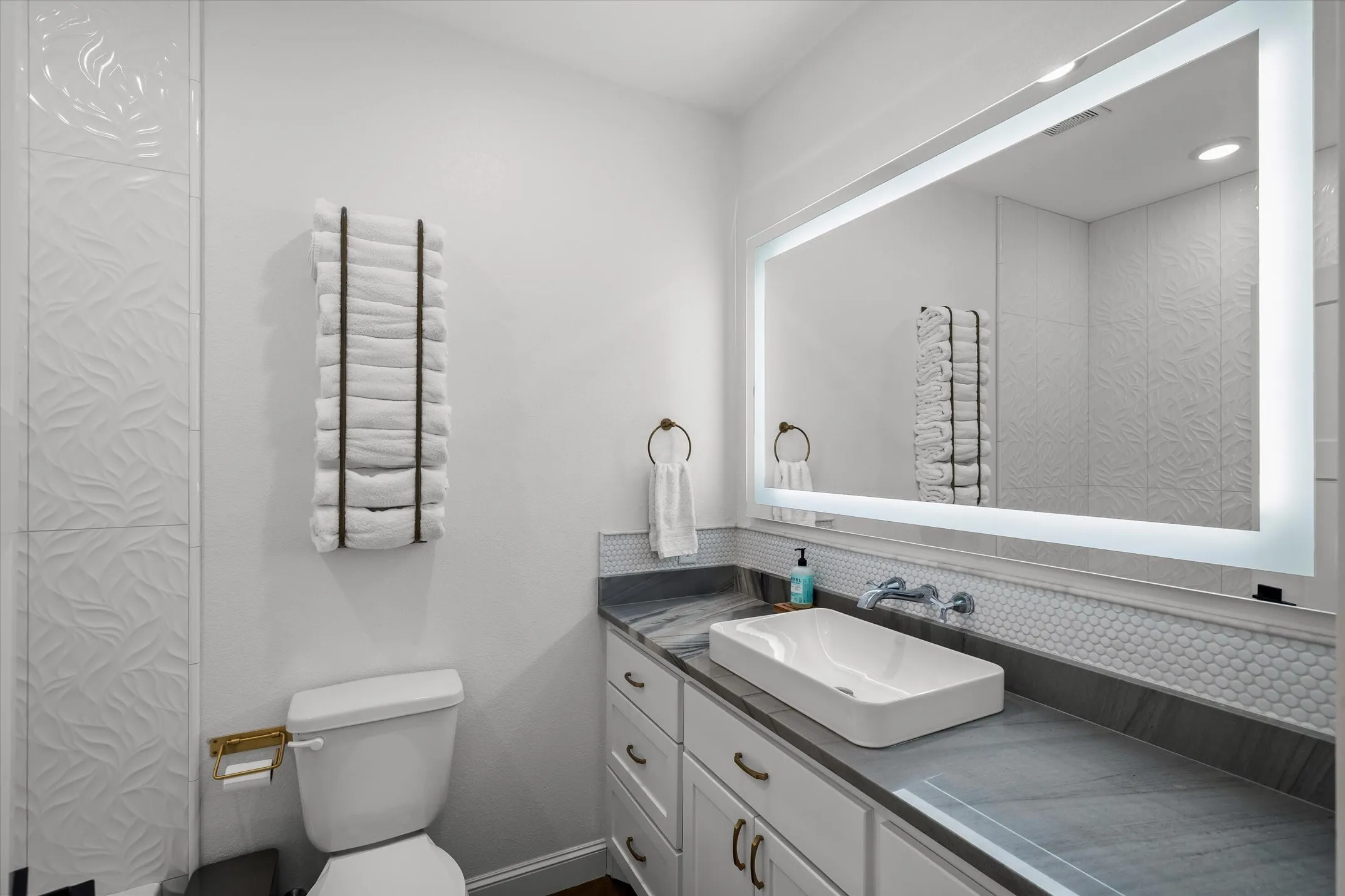 Floating vessel sink with a wall-mounted faucet, illuminated mirror, decorative wall tile, and a vanity with integrated storage